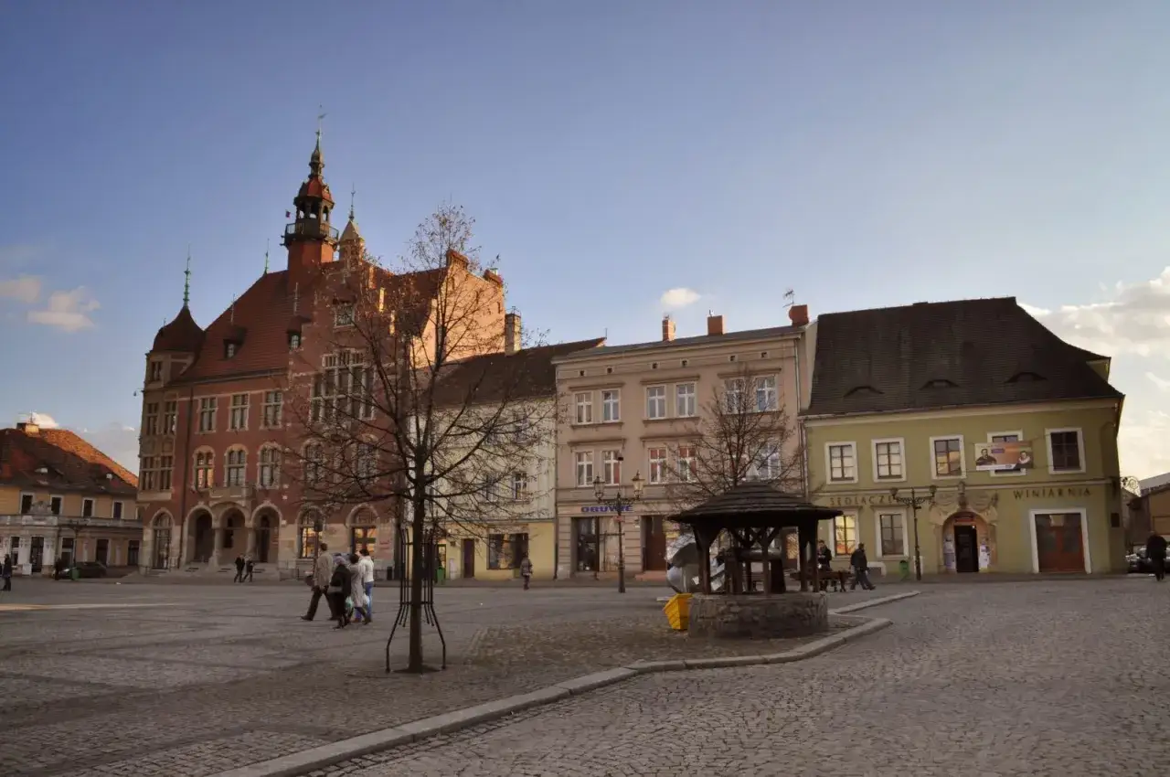 Rynek w Tarnowskich Górach z ratuszem i kolorowymi kamienicami. W centrum studnia i spacerujący ludzie.