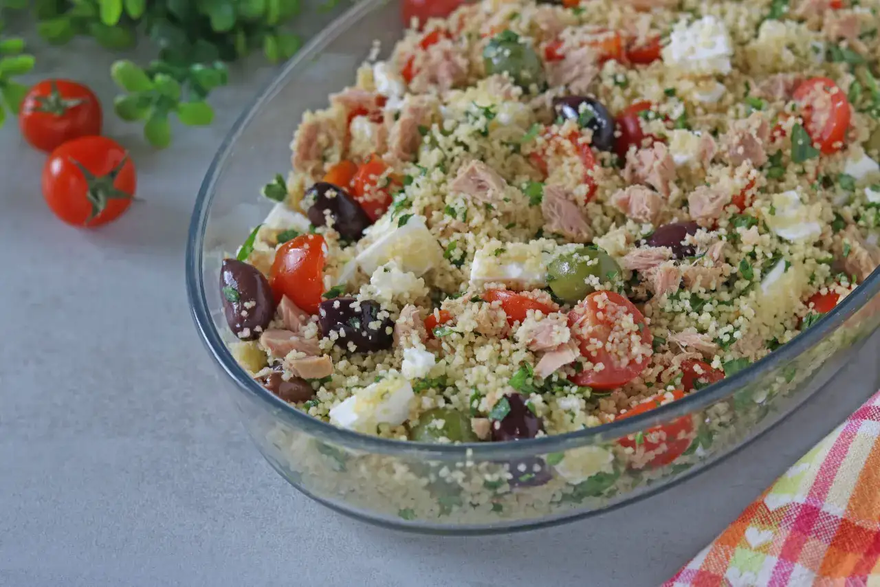 Insalata fresca di cous cous tonno e pomodorini, con olive, feta e prezzemolo, servita in una ciotola di vetro.