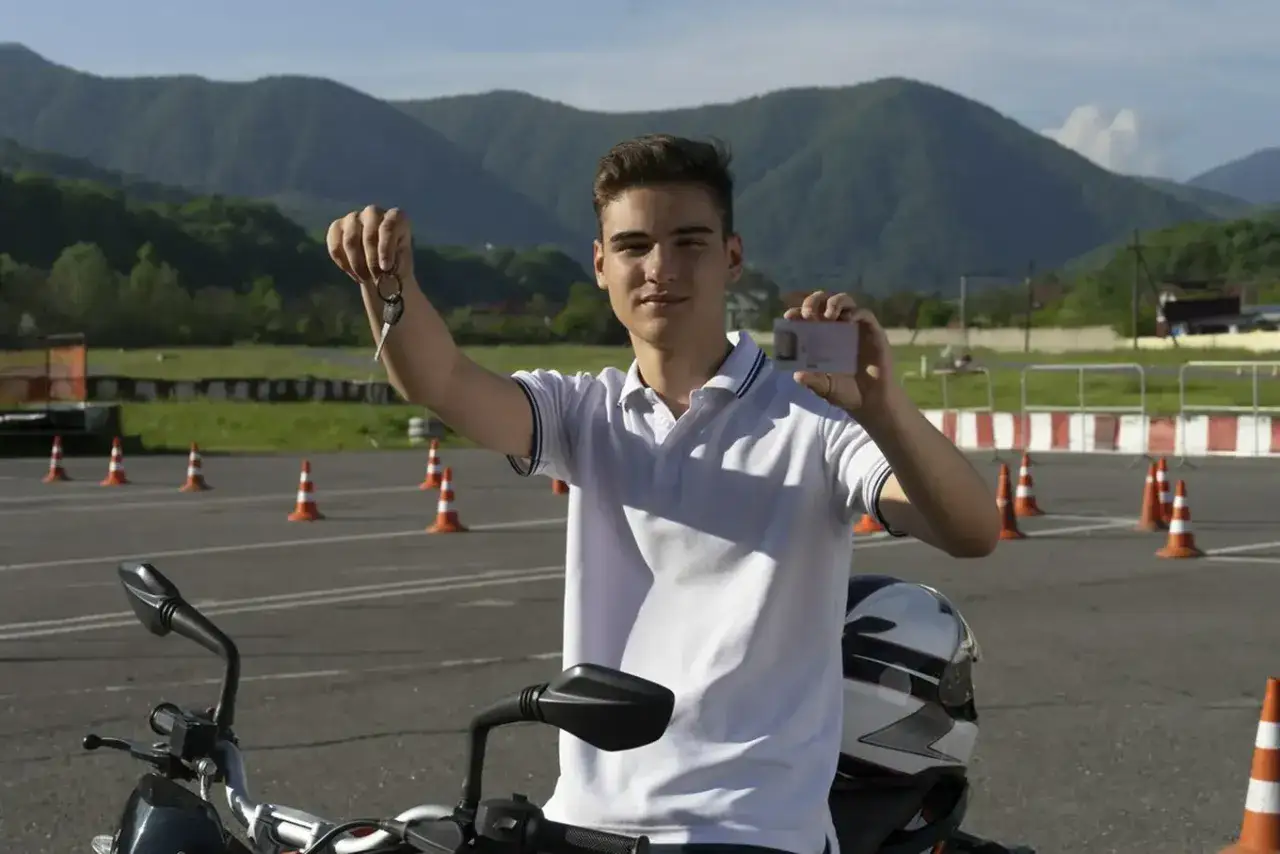 Szczęśliwy młody mężczyzna z kluczykami i prawem jazdy AM za darmo, siedzi na motocyklu na placu manewrowym.