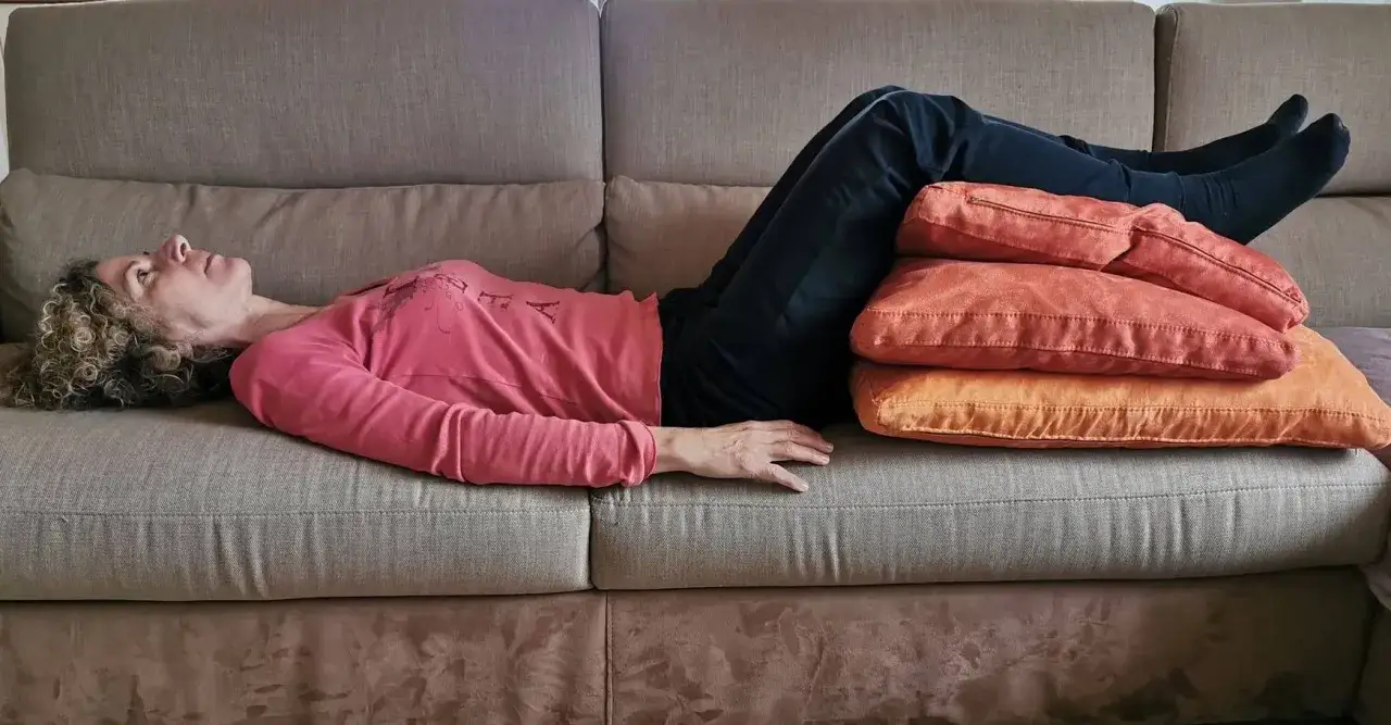 Donna sdraiata sul divano in posizione antalgica, con le gambe sollevate e appoggiate su cuscini arancioni per alleviare il dolore.