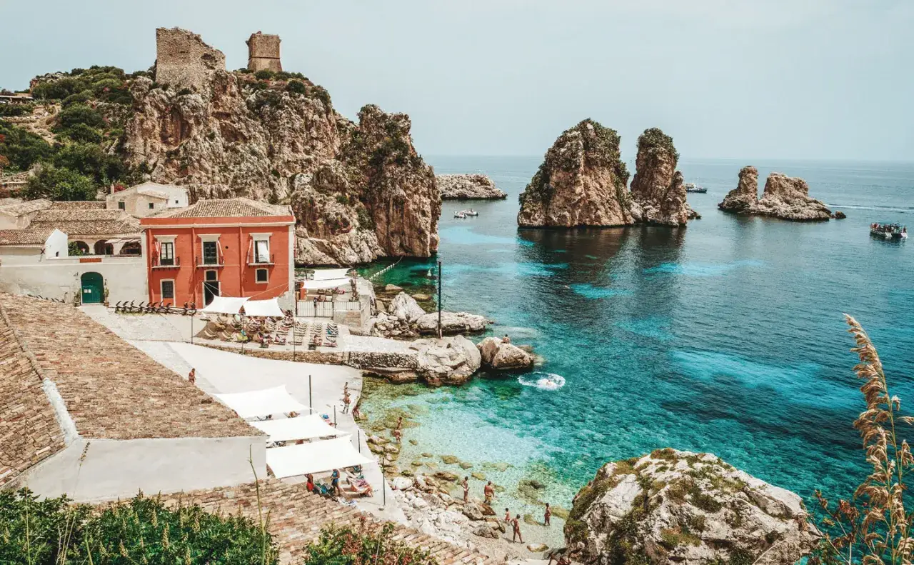 A picturesque coastal village with a red building, rocky outcrops, and turquoise waters, showcasing one of the most beautiful places in Sicily.