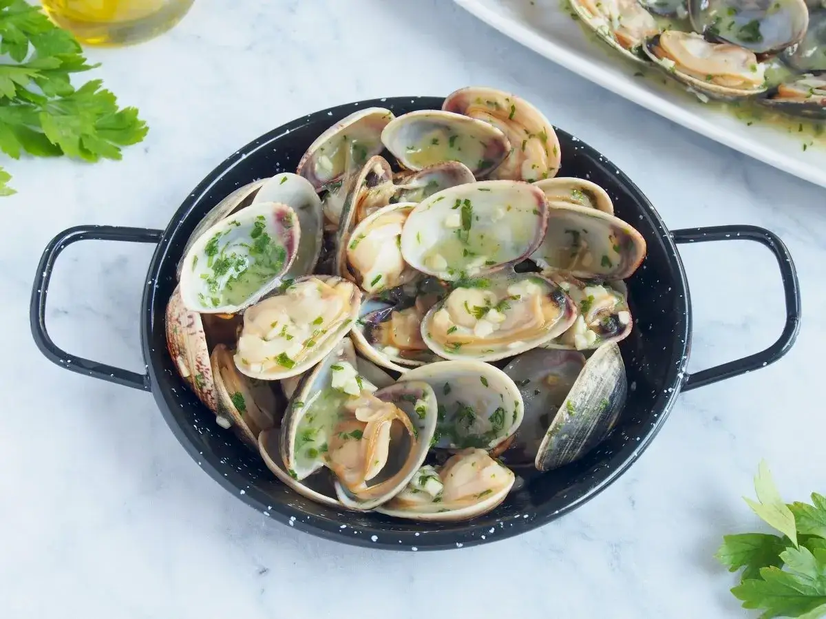 Almejas en salsa verde, con ajo picado y perejil fresco, servidas en una sart&eacute;n negra.
