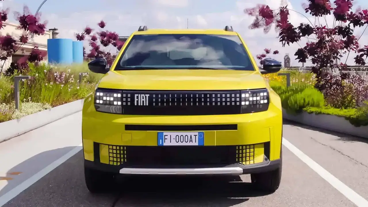 Nowy, żółty Fiat z charakterystycznymi światłami i grillem. W tle kwitnące krzewy i niebieskie cylindry.
