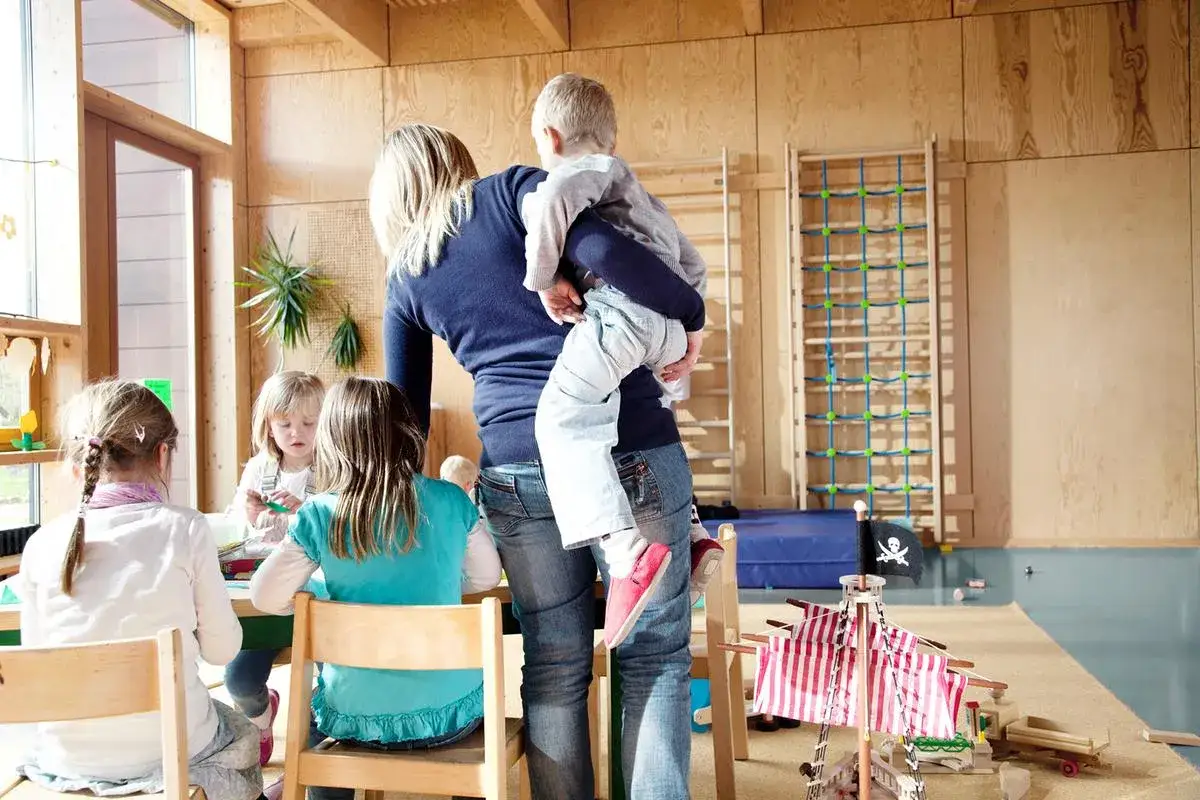 Frau hebt Kind, fragt sich, wieviel kg darf eine Frau heben arbeitsrecht. Kinder spielen im Gruppenraum.