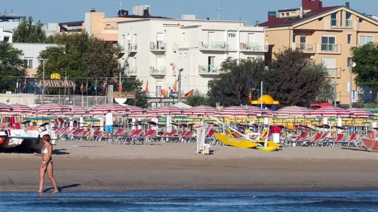 Spiaggia di Rimini Rivazzurra con ombrelloni rossi e bianchi, lettini, una donna in bikini e un bagnino.