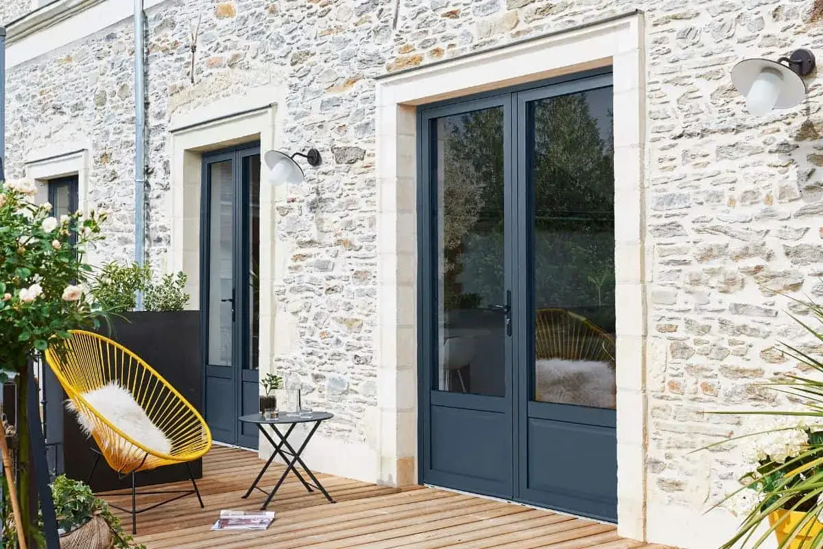 Une porte fen&ecirc;tre qui s'ouvre vers l'ext&eacute;rieur, sur une terrasse en bois, avec une chaise jaune et une table.