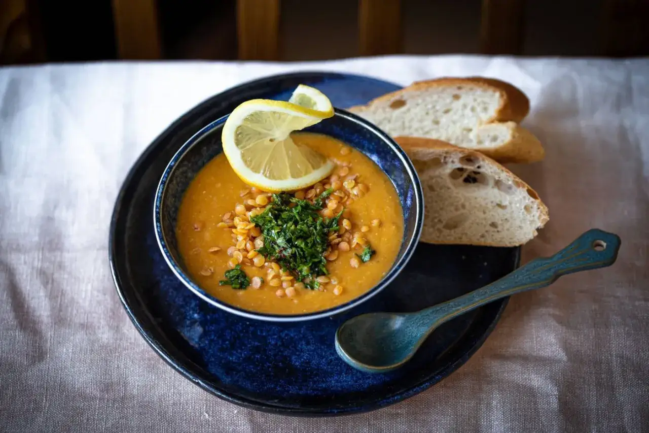 Eine Sch&uuml;ssel mit dampfender, cremiger **roter Linsensuppe**, garniert mit frischer Petersilie und Zitronenspalte, daneben Brot und ein L&ouml;ffel.