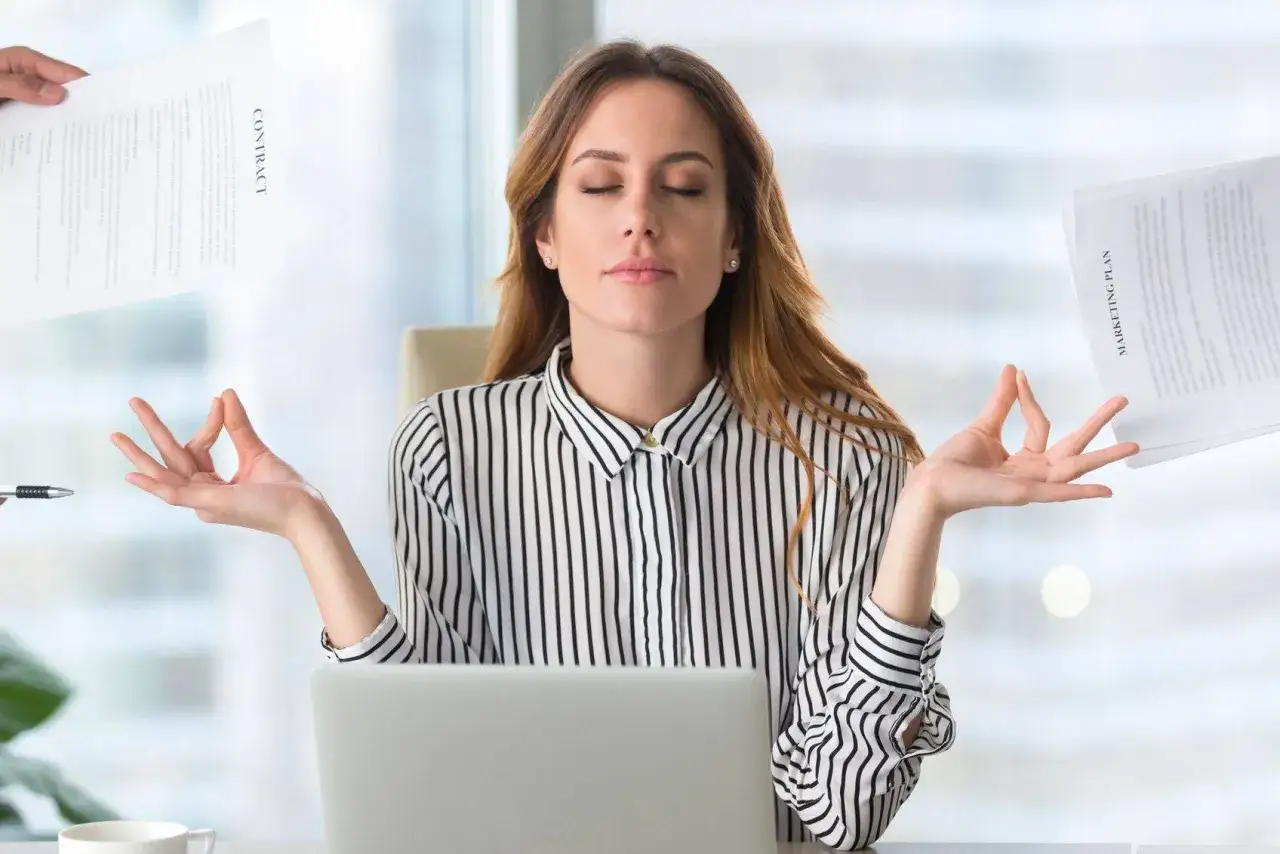 Femme au bureau, les yeux fermés, mains en mudra, entourée de documents. Elle pratique un mantra positif travail pour rester sereine.
