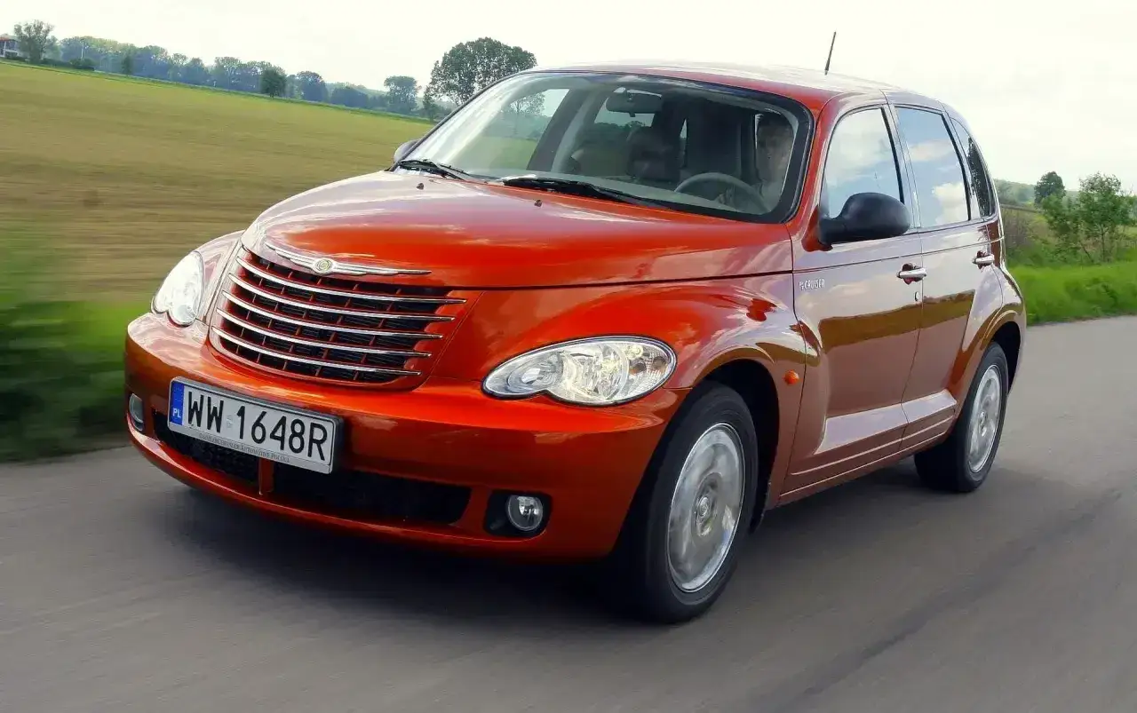 Pomarańczowy Chrysler PT Cruiser jedzie po drodze, za nim zielone pola i drzewa.