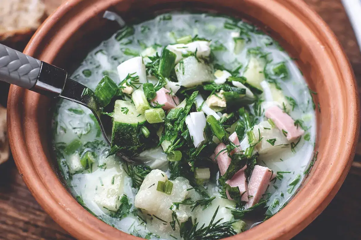 Une cuillère prélève une portion généreuse de cette soupe froide rafraîchissante, garnie de concombre, d'œufs durs, d'herbes fraîches et de jambon.