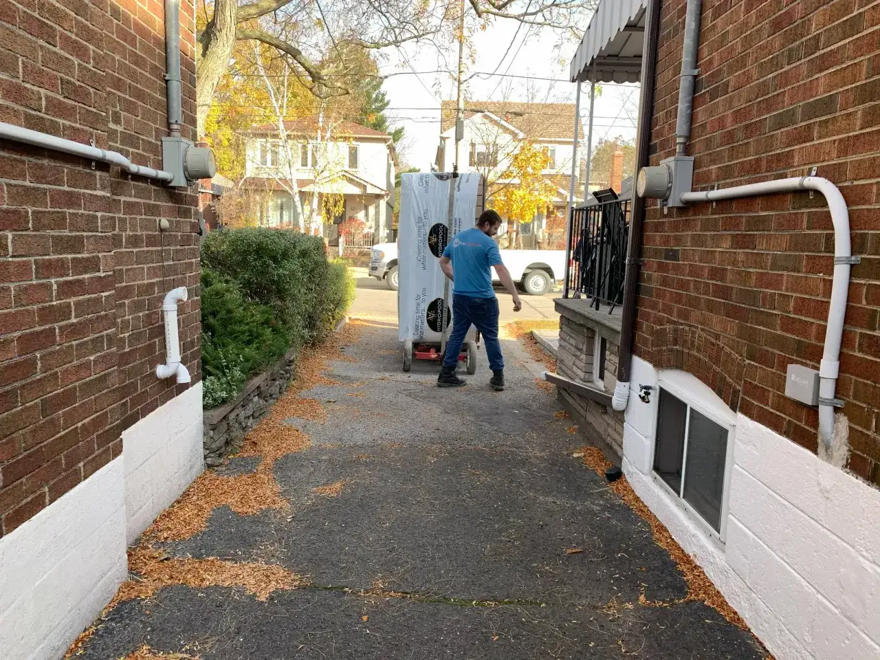 A man is moving a large, wrapped item on a dolly, likely for a hot tub installation, down a narrow driveway between two brick houses.