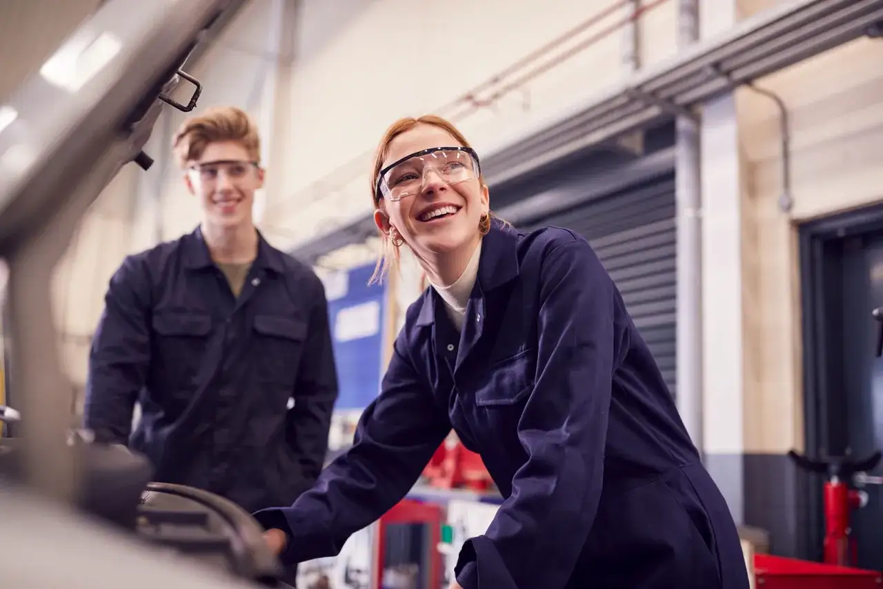 Zwei junge Leute in Blaumann und Schutzbrille arbeiten an einem Auto. Sie machen ein Praktikum ohne Studium und haben sichtlich Spa&szlig;.