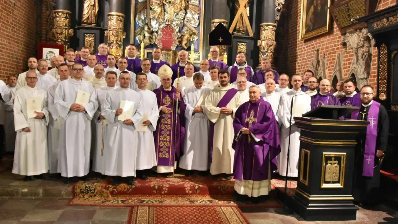 Grupa mężczyzn w szatach liturgicznych, wśród nich akolita w purpurowej dalmatyce, pozuje do zdjęcia w kościele.