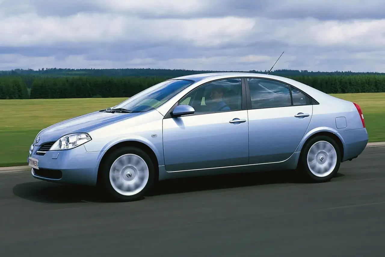 Srebrny Nissan Primera 2002 jedzie po drodze, za nim zielone pola i las.