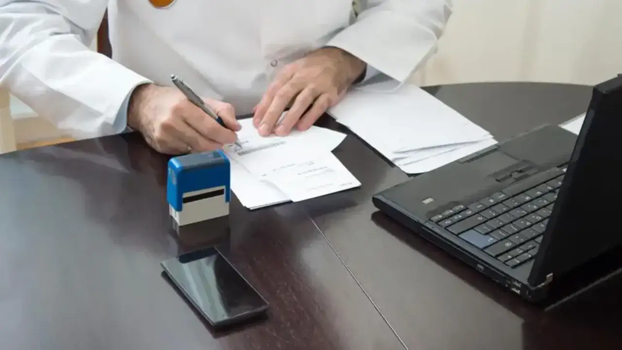 Lekarz wypełnia dokumenty, które mogą być podstawą do ubiegania się o zasiłek chorobowy. Na biurku leży laptop, pieczątka i telefon.