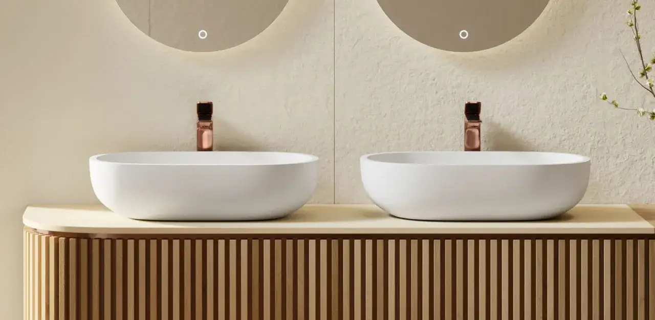 Dos lavabos blancos sobre encimera con grifería cobre. Mueble de baño con lamas de madera.
