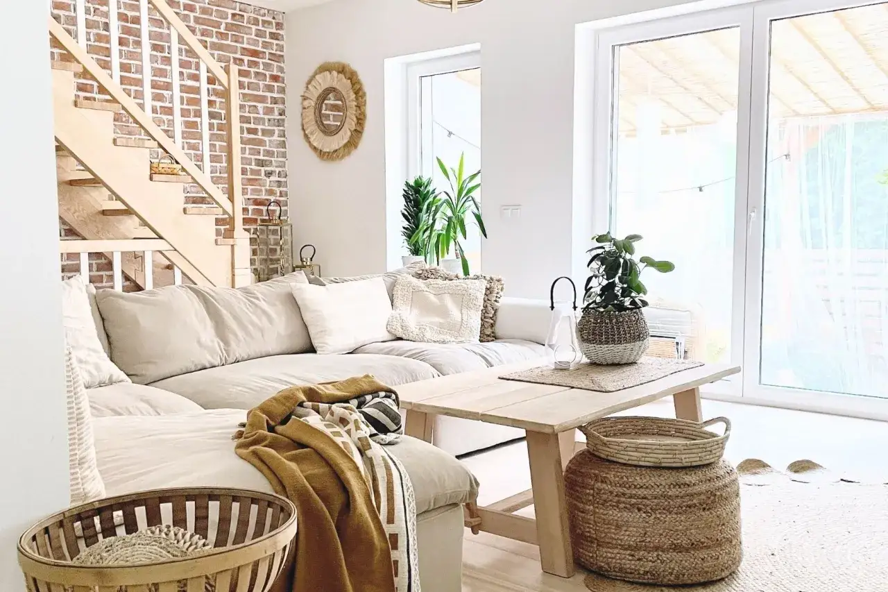 Przytulny salon w stylu scandi boho. Jasna kanapa, drewniane schody z ceglaną ścianą, plecione kosze i rośliny tworzą ciepłą atmosferę.