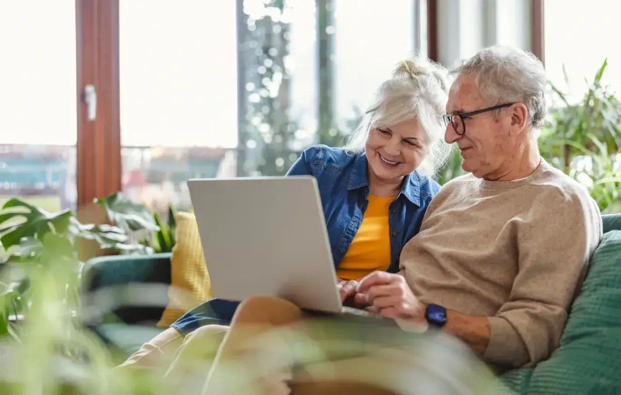 Coppia anziana sorridente guarda un laptop, esplorando le possibilità della domotica per anziani per una vita più semplice e sicura.