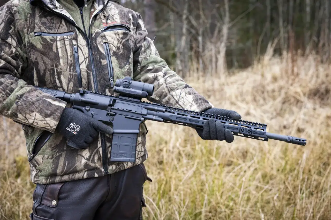 Person i kamouflagekläder håller ett AR-15 gevär med kikarsikte, redo för ar 15 jakt i skogen.