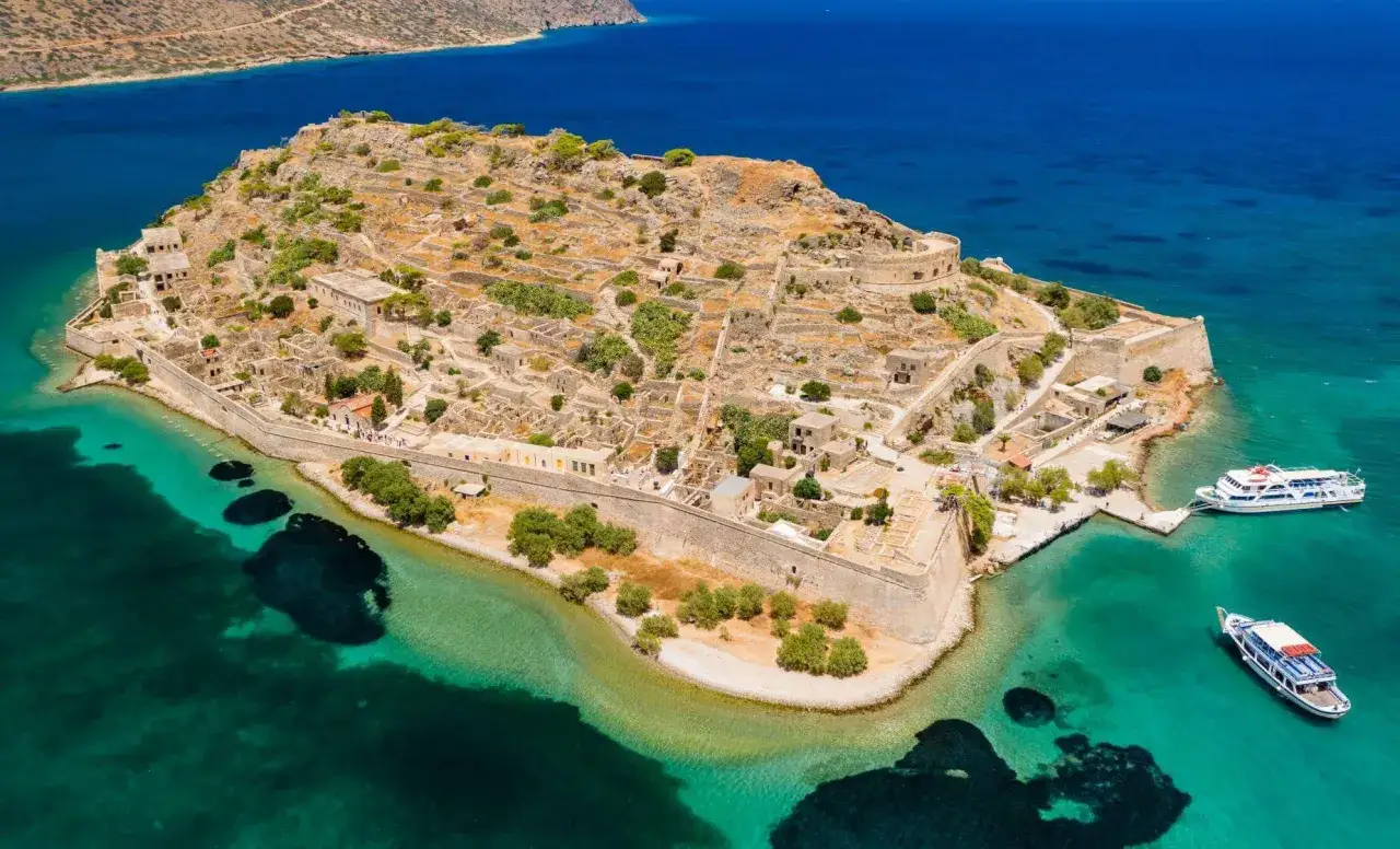 Wyspa Spinalonga z ruinami i turkusową wodą. Dwa statki wycieczkowe zacumowane przy brzegu, idealne na wycieczkę na plażę Vai.