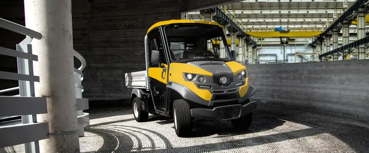 Żółty, kompaktowy polski samochód elektryczny z otwartą paką, zaparkowany w industrialnym otoczeniu.