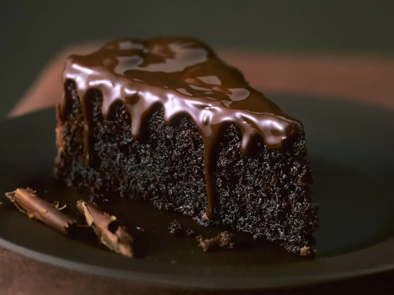 Ein St&uuml;ck saftiger Schoko Kuchen mit gl&auml;nzender Schokoladenglasur, die &uuml;ber den Rand tropft. Daneben liegen zwei Schokost&uuml;ckchen.