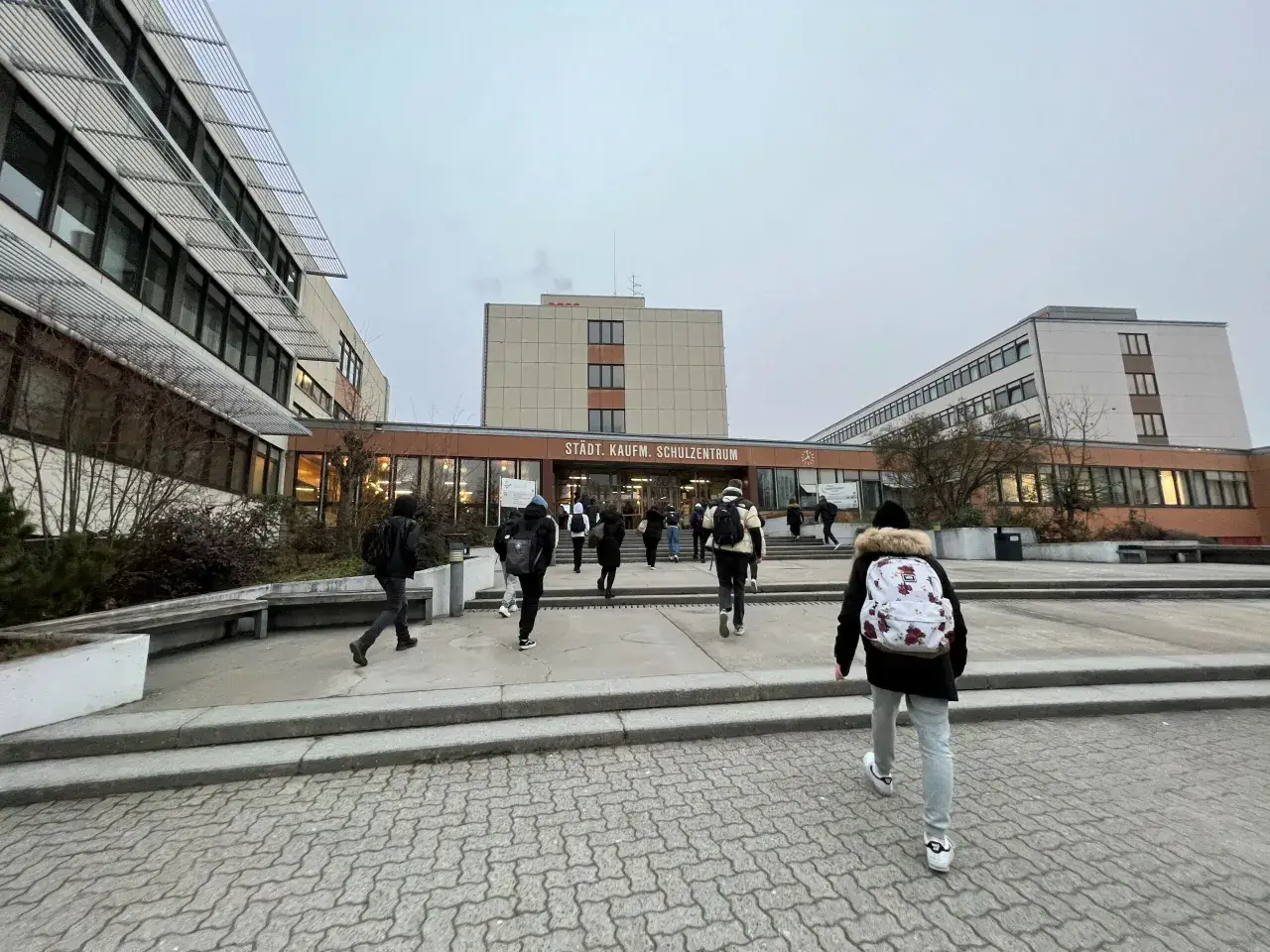 Sch&uuml;ler betreten das St&auml;dtische Kaufm&auml;nnische Schulzentrum. Viele von ihnen tragen Rucks&auml;cke, was auf ein aktives Leben hindeutet, vielleicht sogar auf ein **Fernstudium neben Ausbildung**.