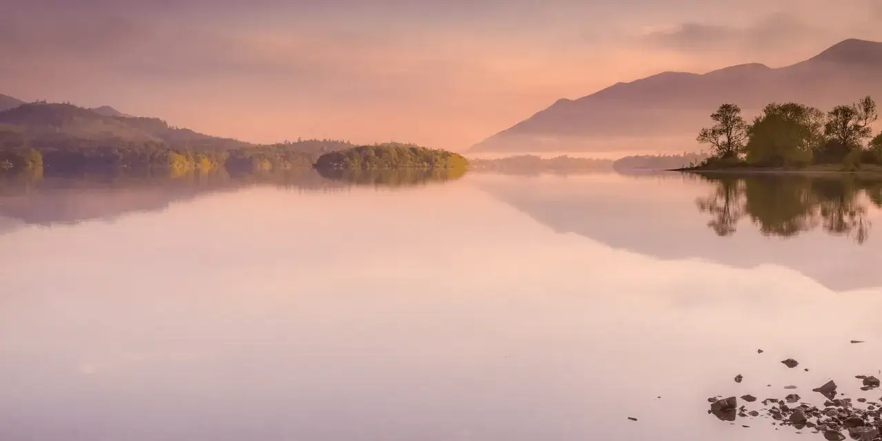 Sanfte H&uuml;gel und B&auml;ume spiegeln sich in einem ruhigen See. Die Szene strahlt eine tiefe Ruhe aus, als ob die Natur **in sich selbst ruhen** w&uuml;rde.