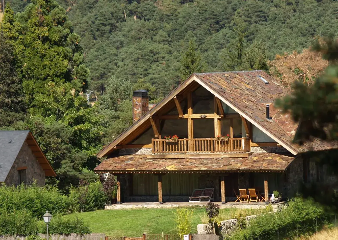 Casas tradicionales de montaña con tejados de pizarra y balcones de madera, rodeadas de exuberante vegetación y un bosque de pinos.