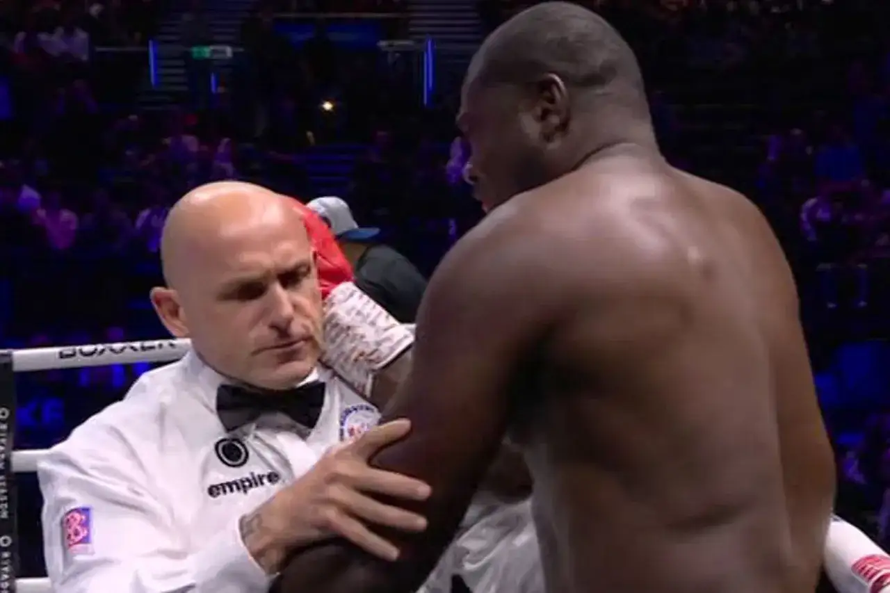 A referee in a white shirt and bow tie holds a boxer's arm, perhaps giving a final instruction before the next round, using boxing terminology.