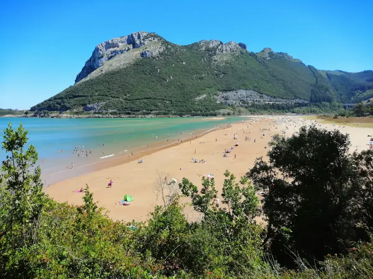 Día soleado en la playa de Sonabia, con gente disfrutando del mar y la arena dorada bajo una imponente montaña verde.