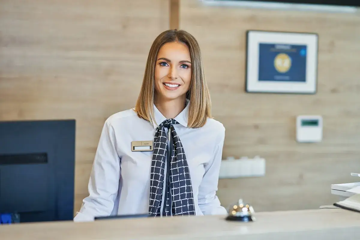 Sorriso accogliente di un addetto al front office in un hotel, con un computer e una campanella sul bancone.