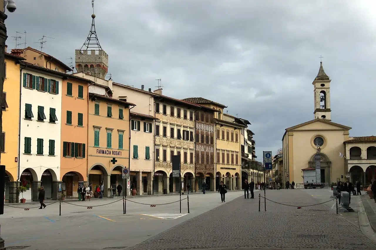 Piazza a Figline Valdarno con edifici storici colorati, una chiesa con campanile e persone che passeggiano sotto un cielo nuvoloso.