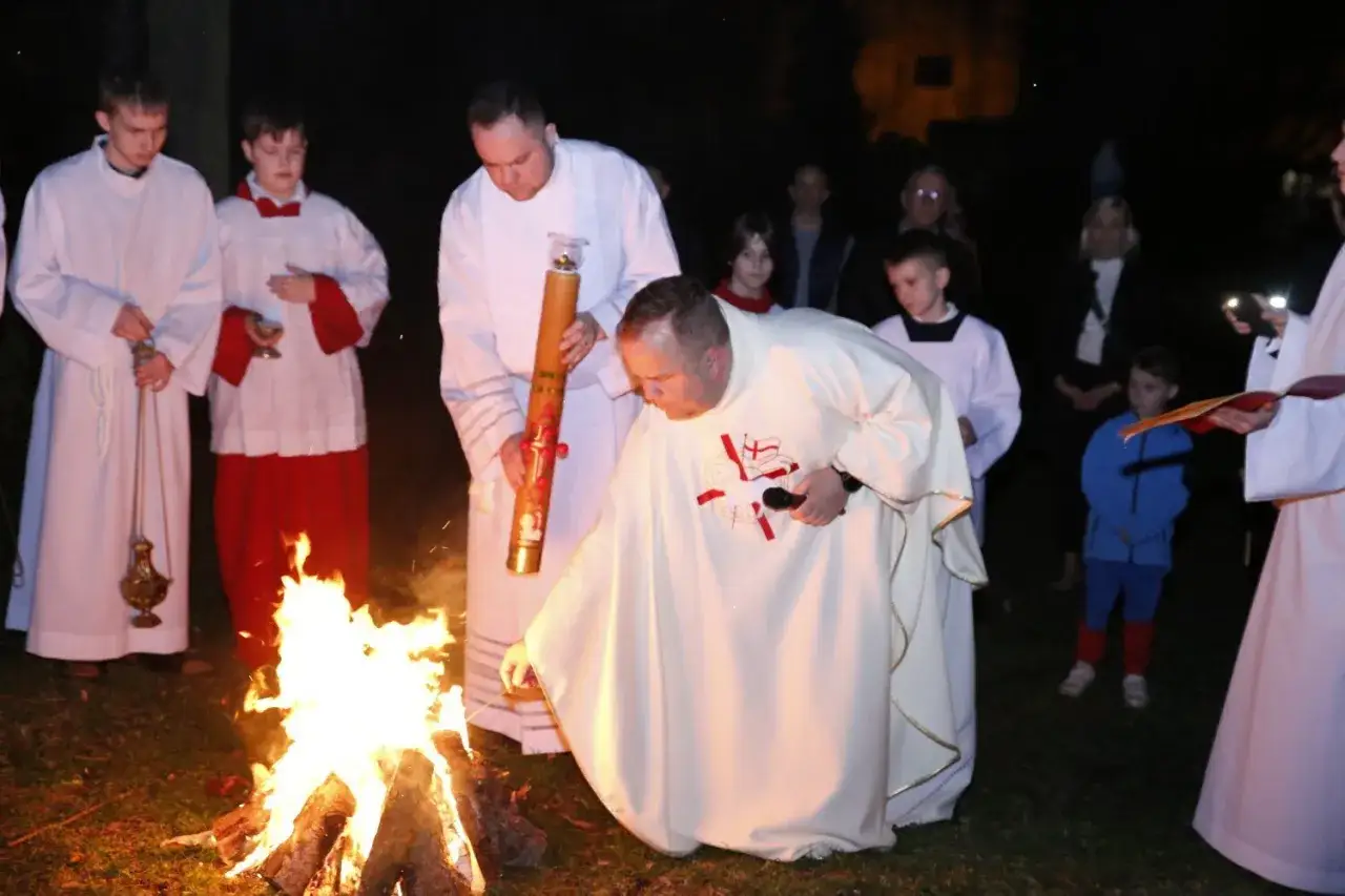Ksiądz dokonuje święcenia wody w kościele przy ognisku. Widać paschalną świecę i ministrant&oacute;w.