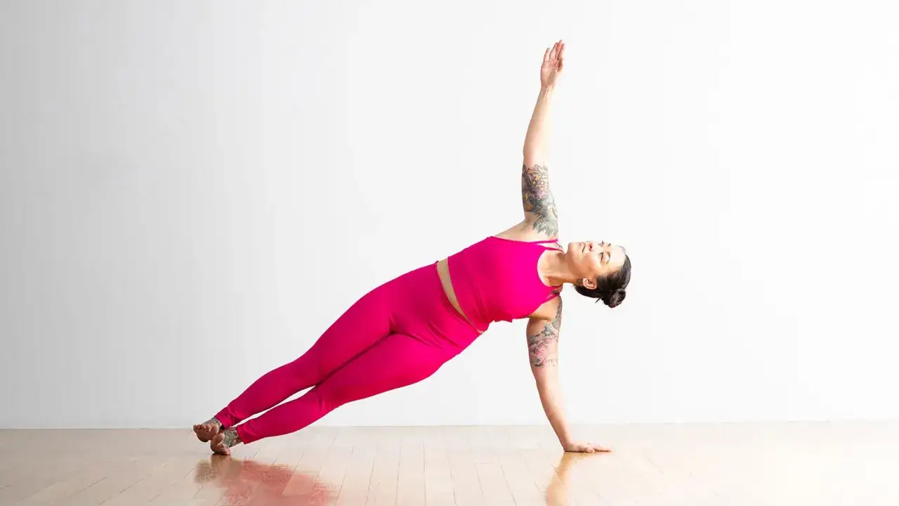 Mujer en plancha lateral yoga, con su brazo derecho extendido hacia el techo y el izquierdo apoyado en el suelo.