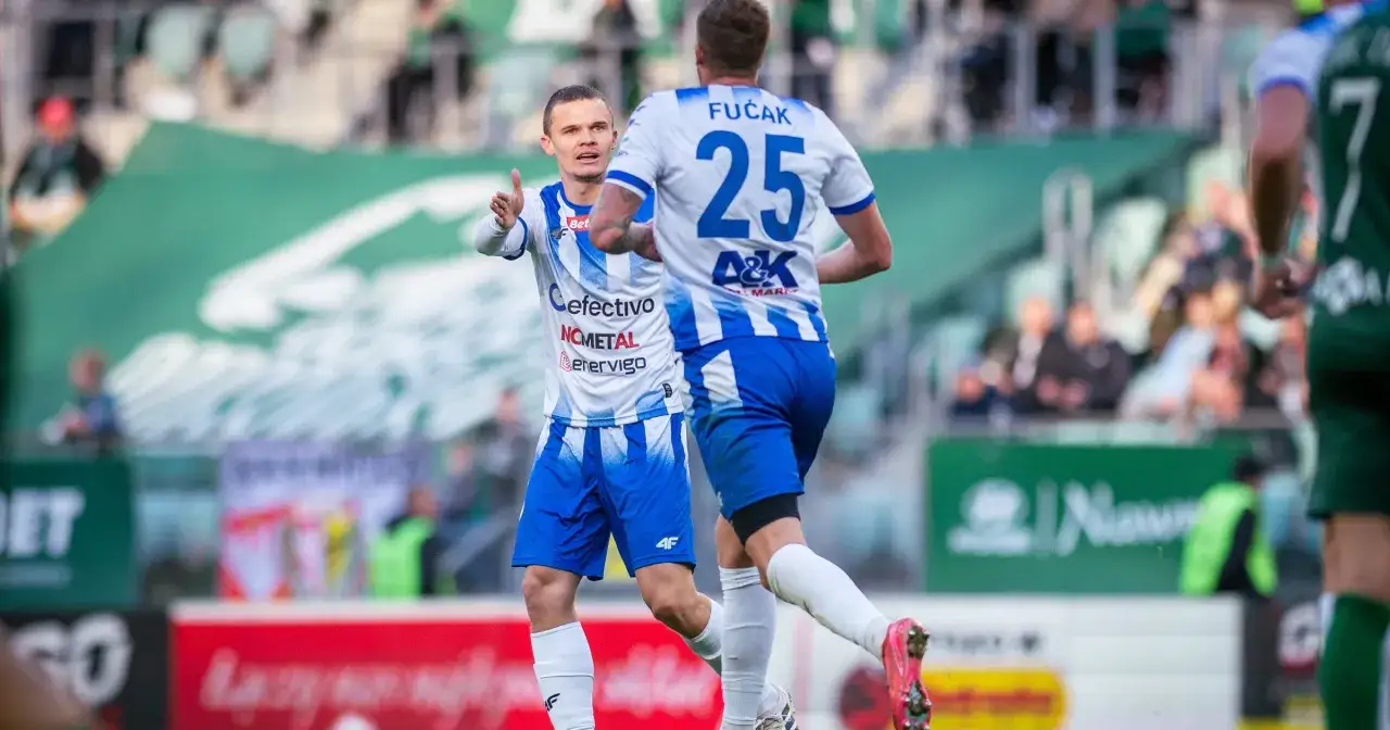 Piłkarze Stali Mielec w akcji. Jeden z nich, w białej koszulce z niebieskimi pasami, gestykuluje do kolegi. Przebieg: Stal Mielec – Miedź Legnica.