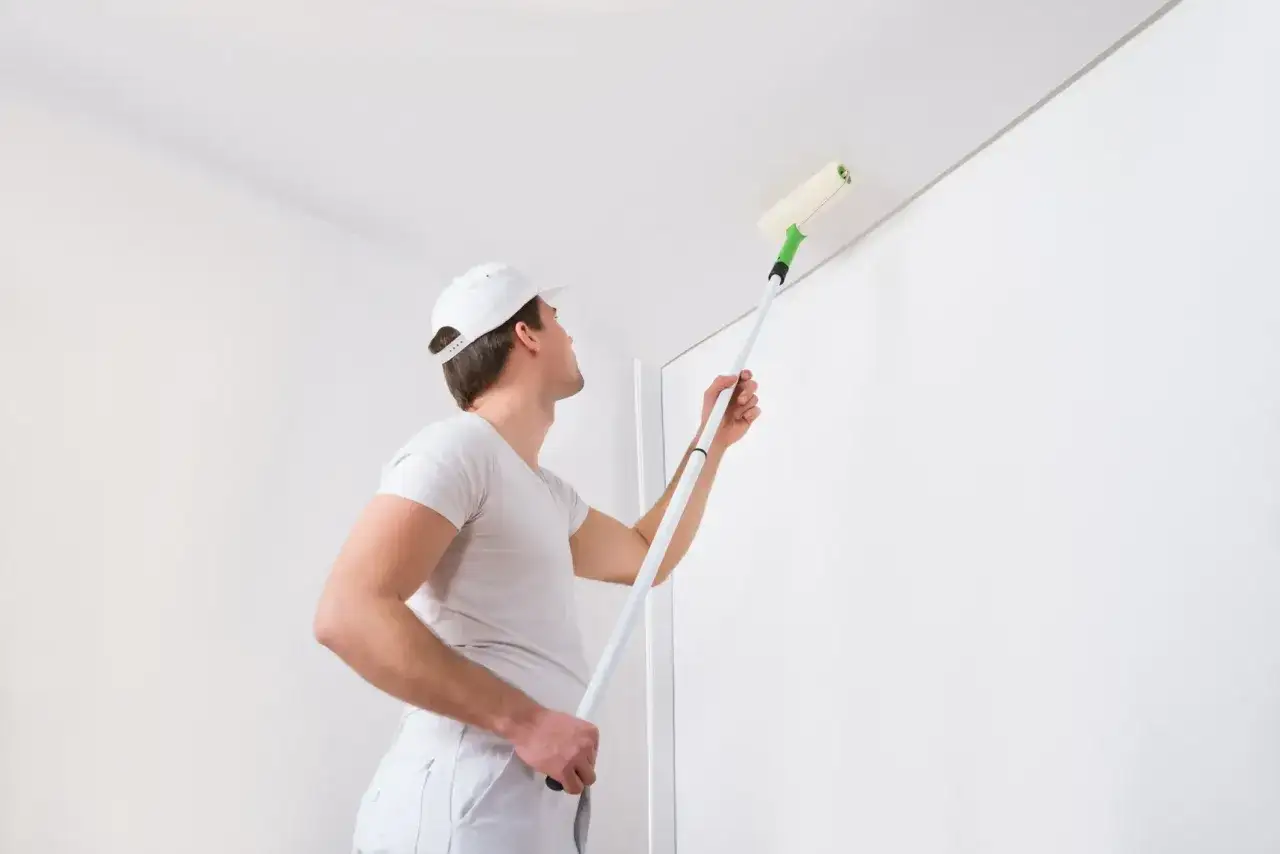 Mann mit Malerhut tapeziert die Decke mit einer Farbrolle.