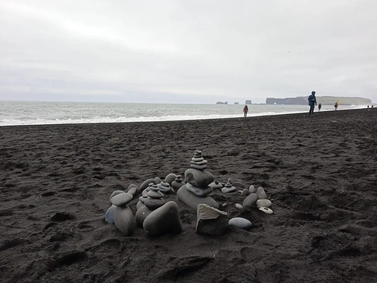 Kamienne stosy na czarnej plaży Reynisfjara, z widokiem na wzburzone morze i skaliste wybrzeże w oddali.