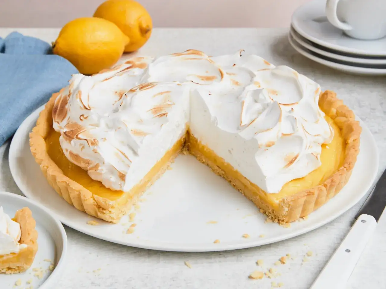 Eine köstliche Tarte au Citron mit fluffiger Baiserhaube, daneben frische Zitronen und eine blaue Serviette.