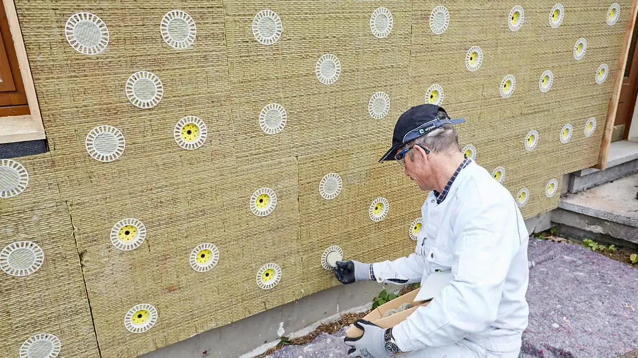 Pracownik montuje kołki do styropianu na ocieplonej ścianie budynku.