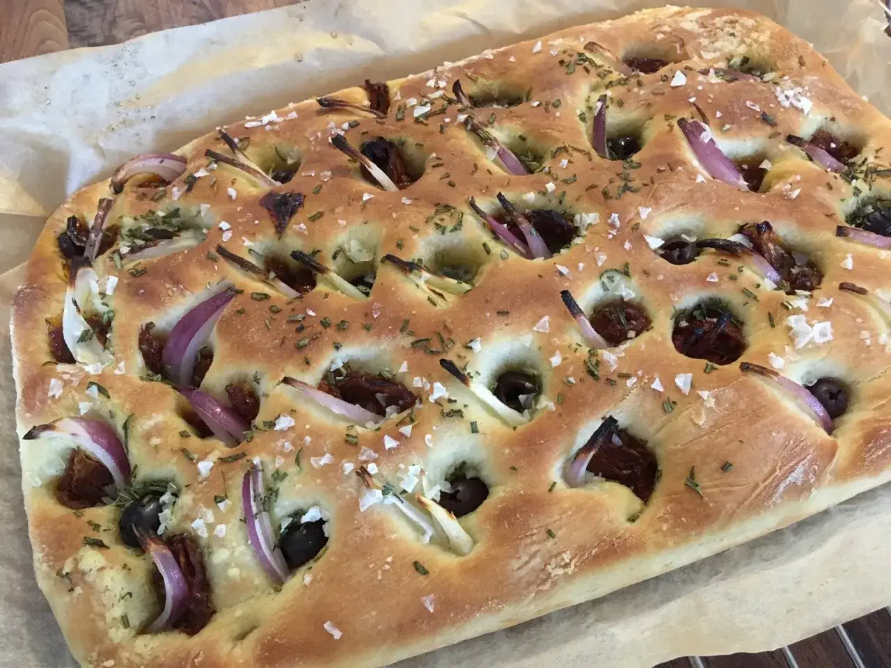 En gyllene focaccia långpanna med rödlök, oliver och soltorkade tomater, strödd med örter och flingsalt.