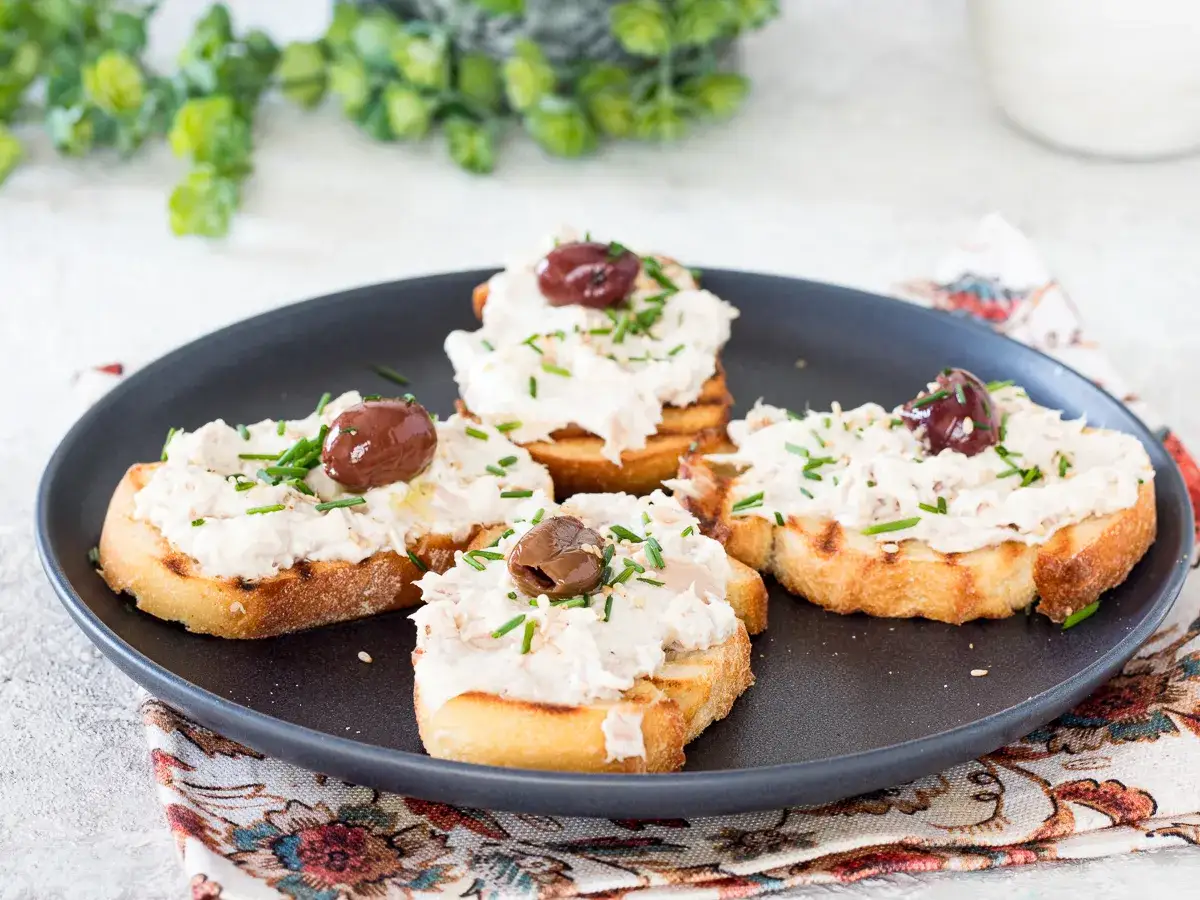 Quattro crostini di pane con crema di pesce, olive e erba cipollina su un piatto scuro.
