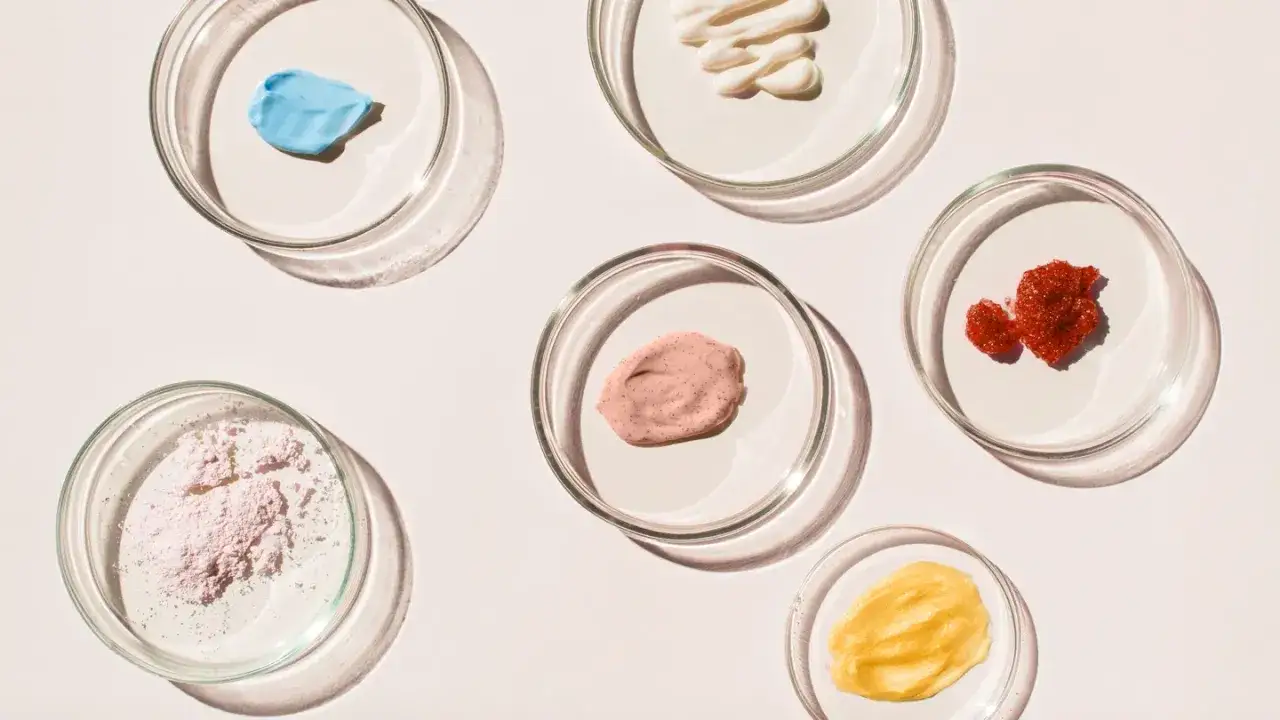 Sechs Petrischalen mit verschiedenen Kosmetikproben: blaue Creme, weißer Balsam, rosa Peeling, roter Granulat, gelbe Creme und rosa Puder. Linalool ist ein Duftstoff, der oft in solchen Produkten vorkommt.