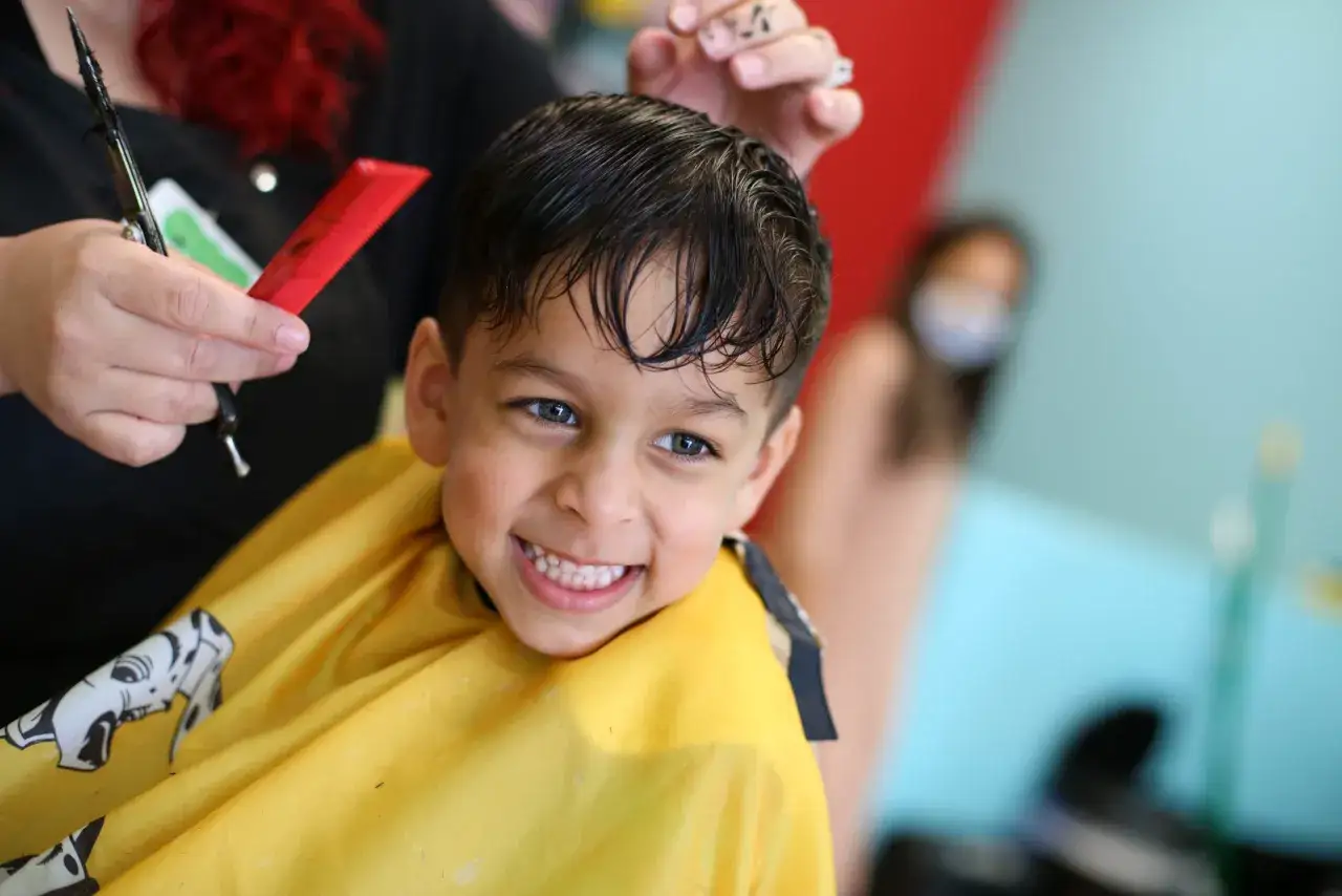 Ein Junge mit einem breiten Lächeln bekommt einen Haarschnitt. Die Friseurin hält eine Schere und einen Kamm bereit, um die **Jungs Frisuren Namen** zu kreieren.