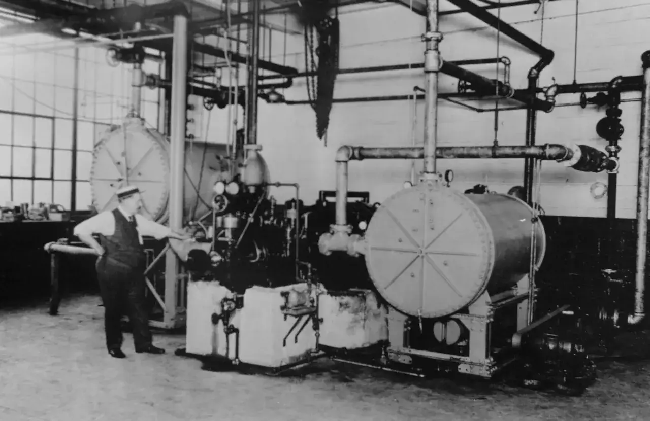 Un hombre con sombrero señala una máquina industrial, quizás la que usó Willis Carrier, quien inventó el aire acondicionado.