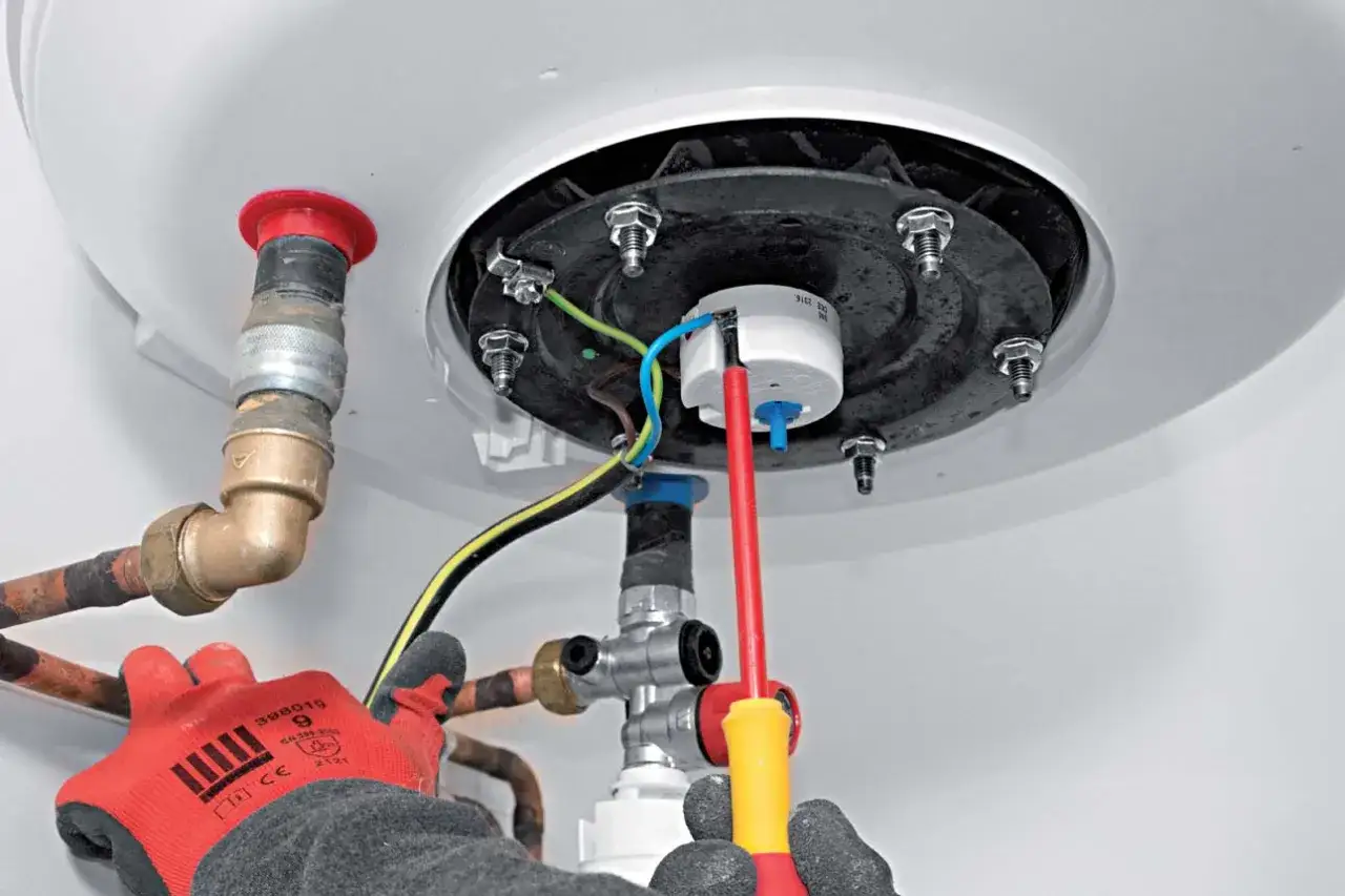 Main d'un technicien en gant rouge effectuant l'installation d'un chauffe-eau, connectant les fils électriques et les tuyaux de cuivre.