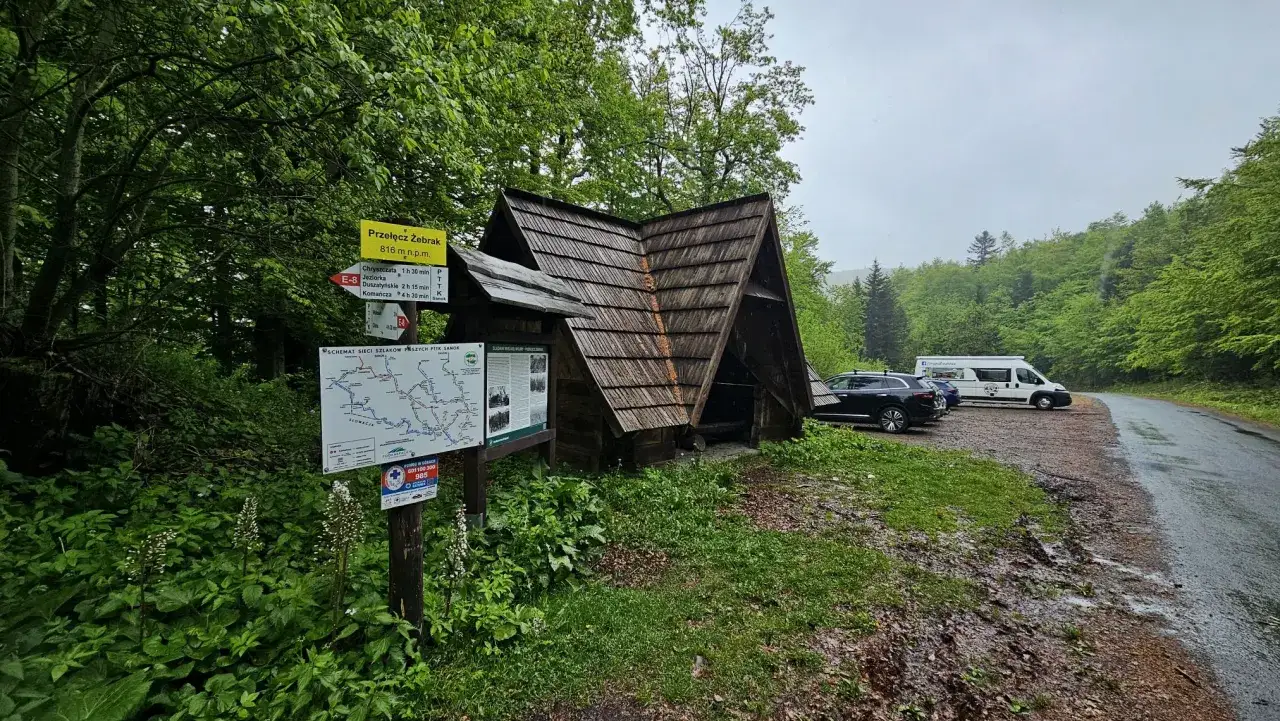 Drewniana wiata na przełęczy Żebrak, obok drogowskazów i mapy szlaków. Zaparkowane samochody i kamper.