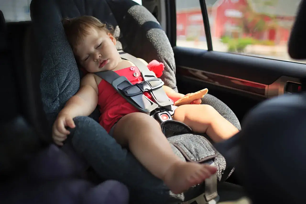Un bébé dort paisiblement dans son siège auto, un biscuit à la main. Le transport enfant voiture est sécurisé.