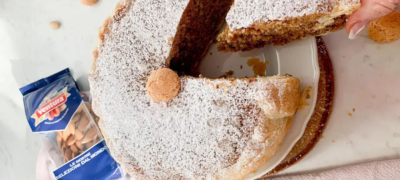 Una fetta di torta russa di Verona, cosparsa di zucchero a velo e decorata con un amarettino. Accanto, una busta di mandorle Ventura.