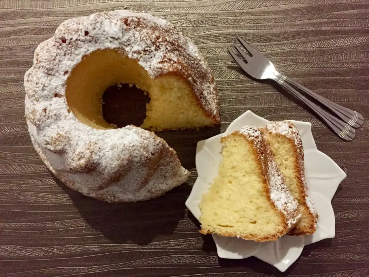 Puszysta babka z majonezem, sprawdzony przepis, oprószona cukrem pudrem. Obok talerzyk z dwoma kawałkami ciasta i widelczyki.